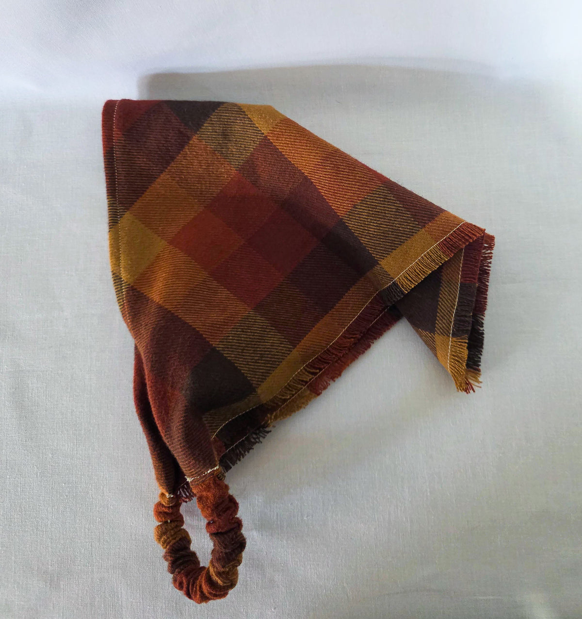 A plaid bandana in warm autumn colors, featuring a fringed edge and an elastic scrunchie loop, laid flat on a light fabric background.