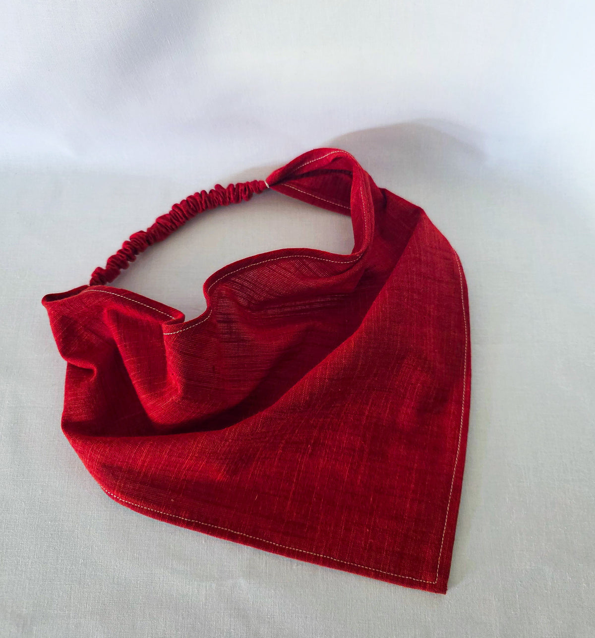 A red triangular hair bandana with an elasticized neck strap, laid flat on a white fabric background.