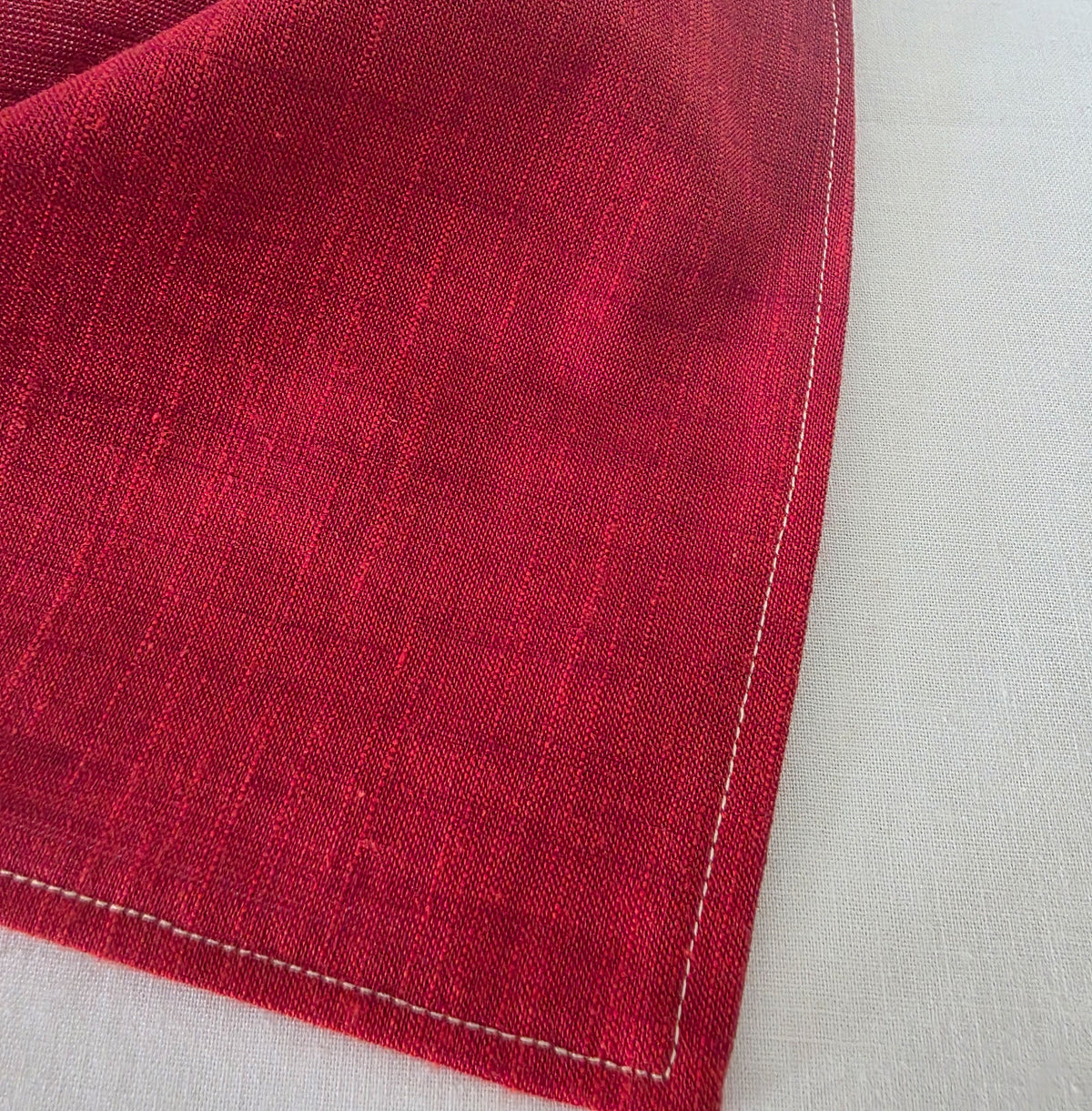 A close-up of a deep red woven hair bandana showing its textured weave and white contrast stitching along the edge, placed on a light beige linen surface.