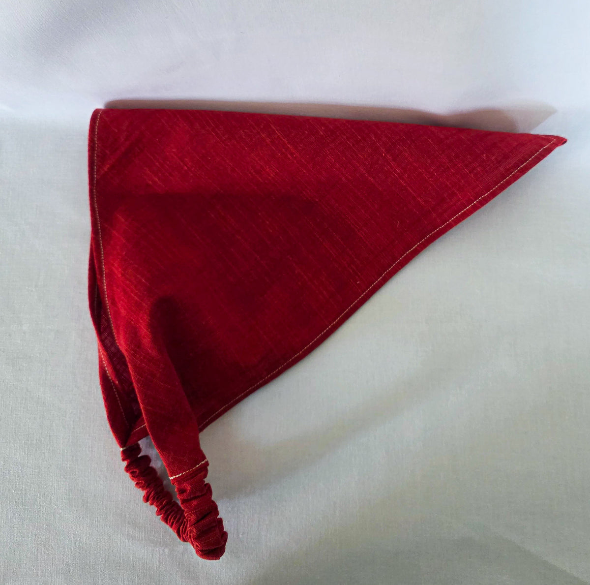 A red triangular bandana with a gathered elastic strap, laid flat on a white fabric background.