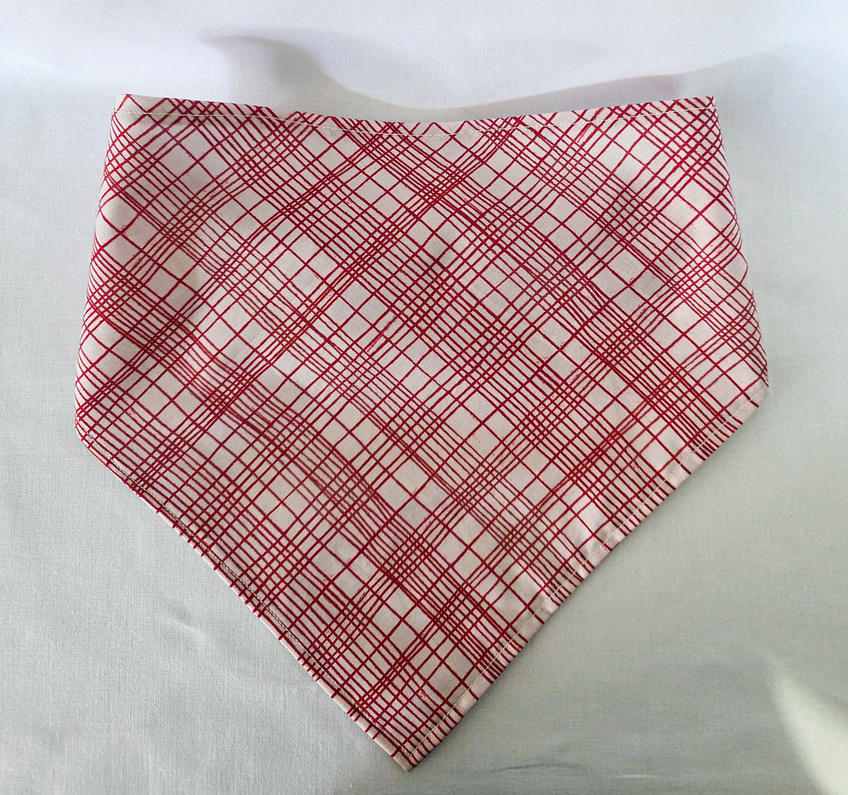 A triangular bandana with a red and white plaid pattern, laid flat on a white fabric background.