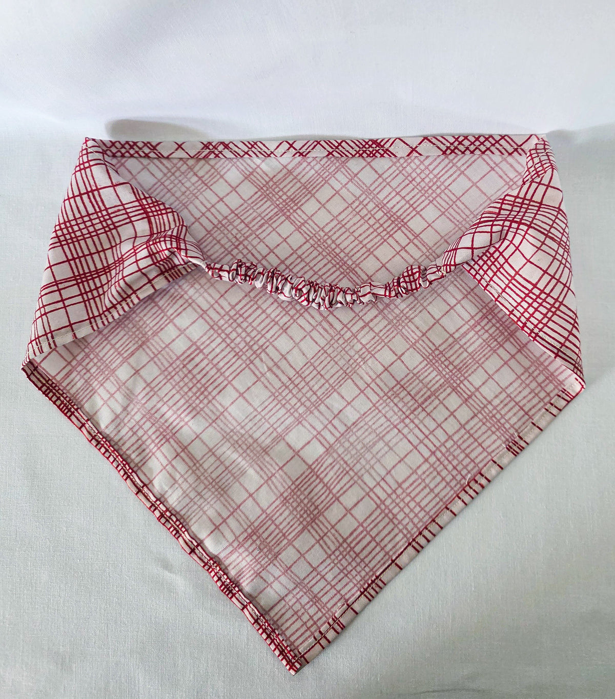 A triangular hair bandana with a red and white plaid pattern, laid flat on a white fabric background.