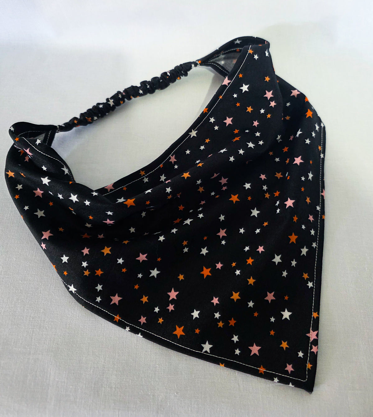 A black triangular hair bandana with an elastic strap, decorated with a pattern of small white, pink, and orange stars, laid flat on a white fabric background.