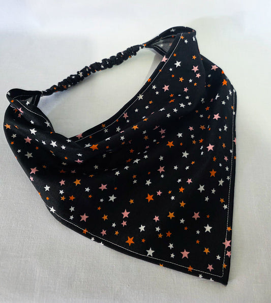 A black triangular hair bandana with an elastic strap, decorated with a pattern of small white, pink, and orange stars, laid flat on a white fabric background.
