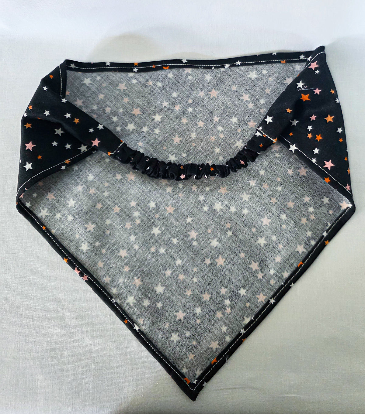 A triangular bandana laid flat on a light fabric surface. It has an elasticized top band and the print is small multicolored star prints including white, pink, orange, 