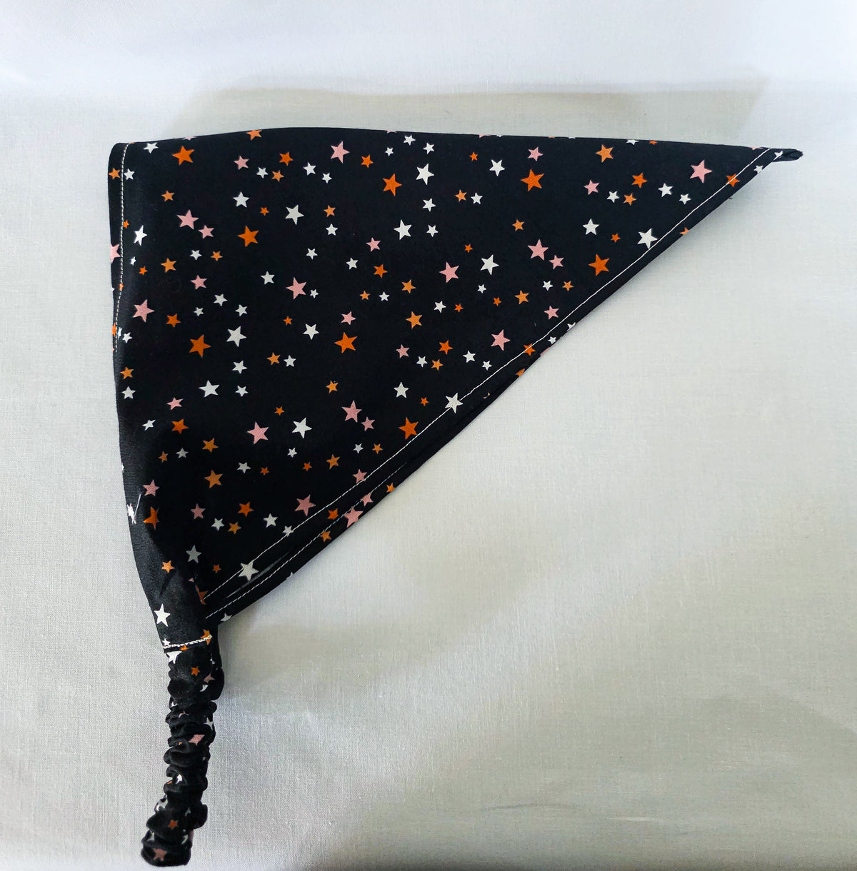 A black hair bandana laid on its side showing the elastic band. The bandana has a scattered star pattern in white, pink, and orange. 