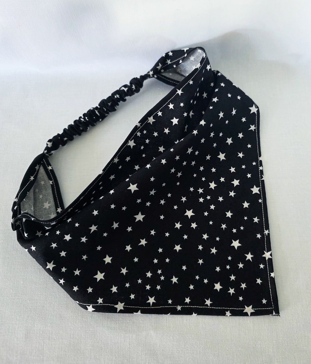 A black triangular hair bandana with a white star pattern, it has an black elastic strap, laid flat on a light gray fabric background. 