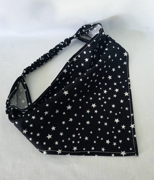 A black triangular hair bandana with a white star pattern, it has an black elastic strap, laid flat on a light gray fabric background. 