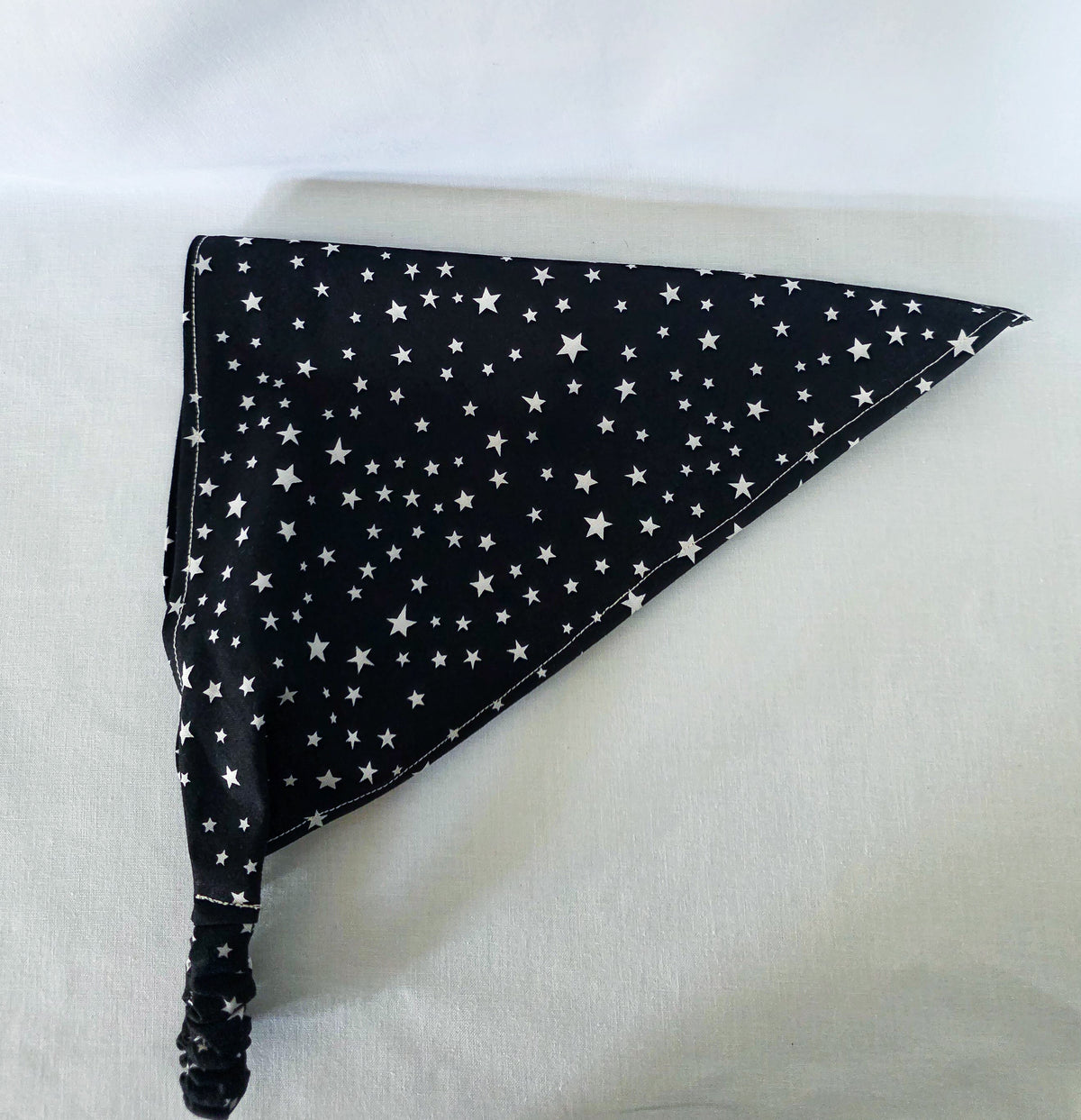 A triangular hair bandana with a black background and white star pattern, it  has an elastic strap, displayed on a light fabric background.