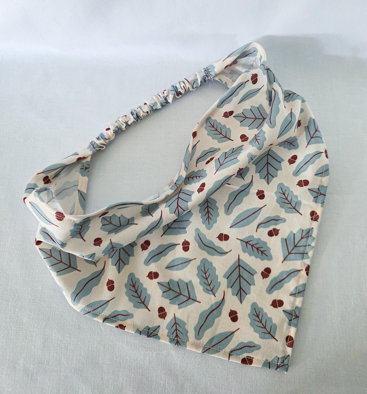 A hair bandana with a gathered elastic strap, featuring an print of blue leaves and small brown acorns on a cream background, laid flat on a light  surface.