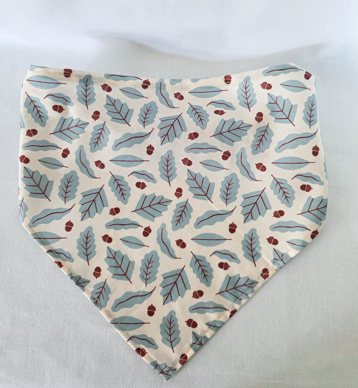 A hair bandana laid flat, featuring an print of blue leaves and small brown acorns on a cream background, on a light  surface.