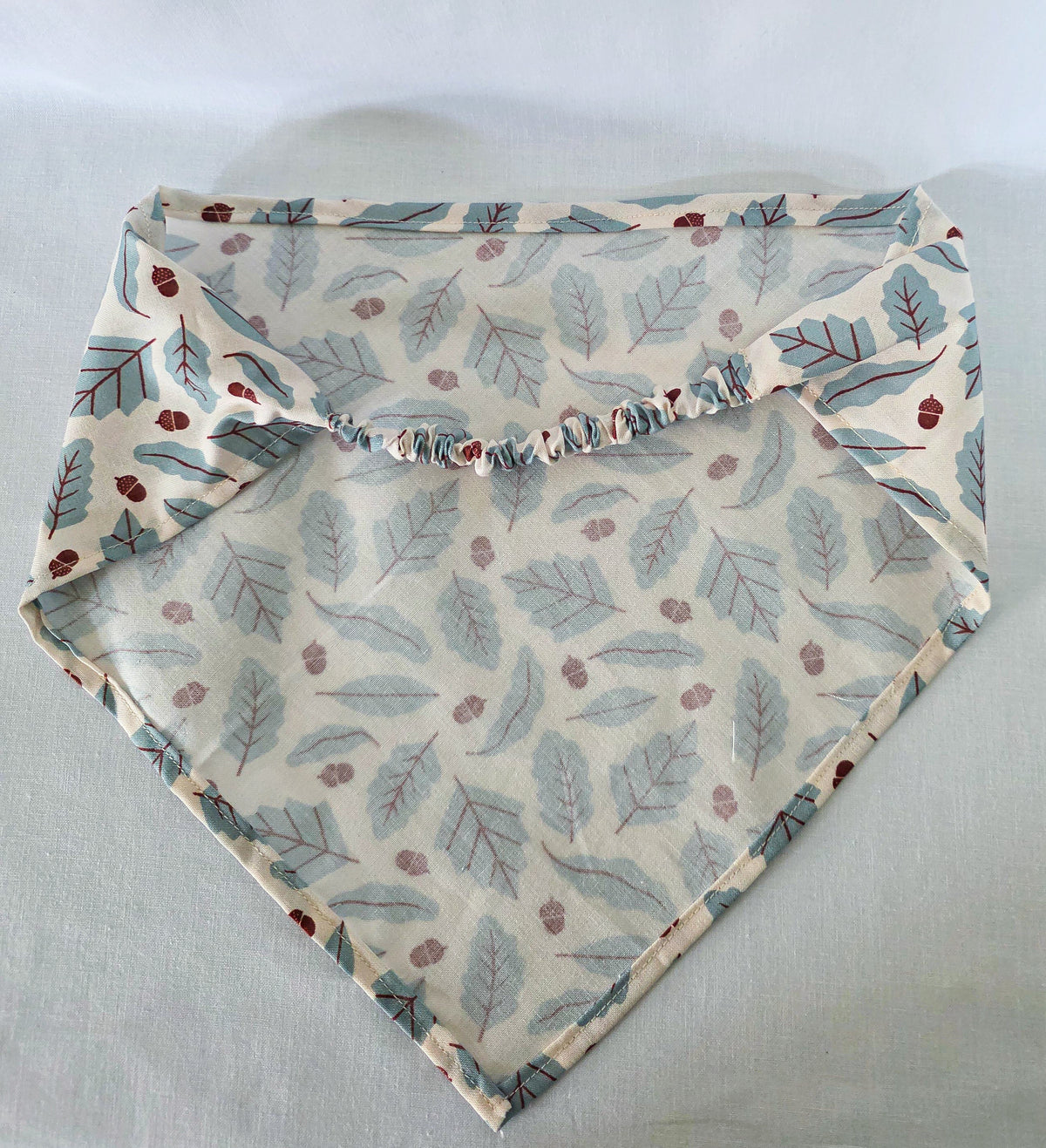 A triangular bandana laid flat. The fabric has an off white base with a repeating pattern of muted blue leaves and small brown acorns, finished with a gathered elastic band.