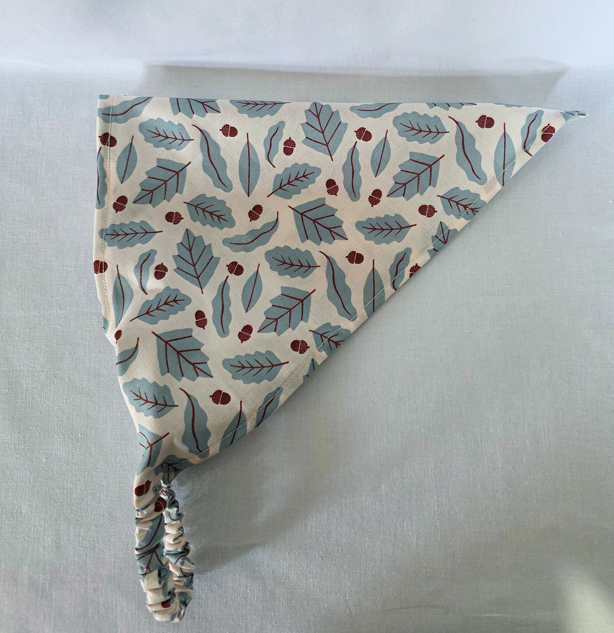 A triangular hair bandana made of cream fabric with a repeating pattern of muted blue leaves and small brown acorns, finished with a gathered elastic strap, laid flat on a light background.