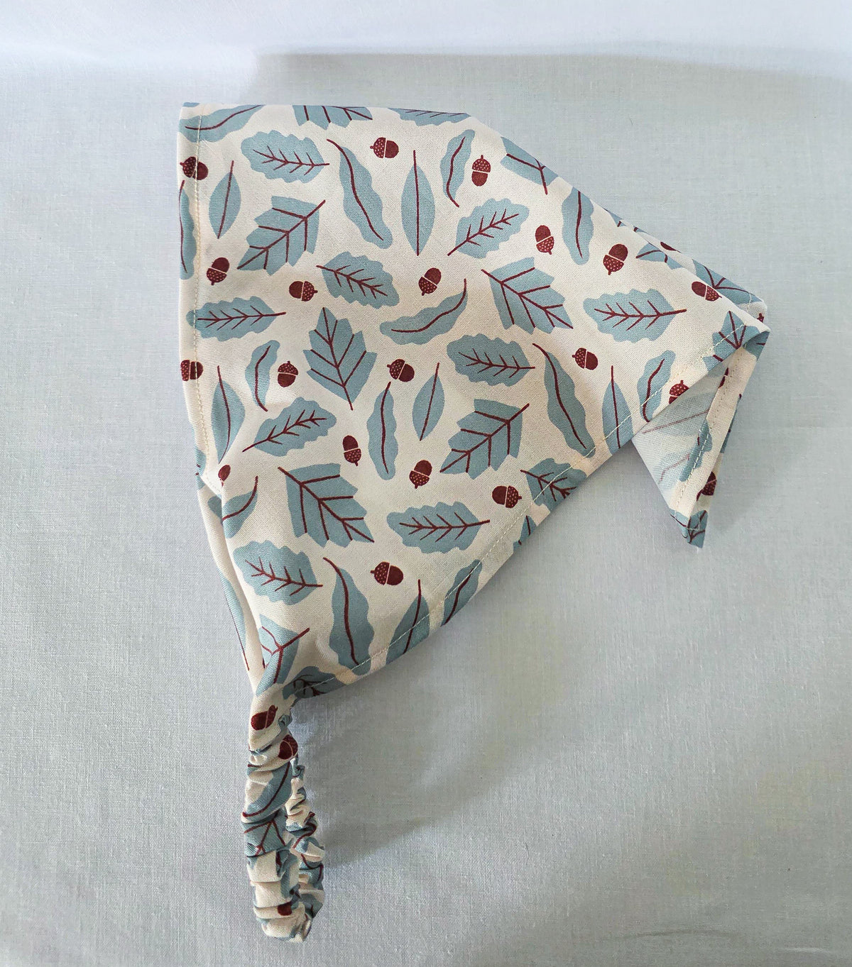 A triangular hair bandana made of cream fabric with a repeating pattern of muted blue leaves and small brown acorns, finished with a gathered elastic strap, laid flat on a light background.