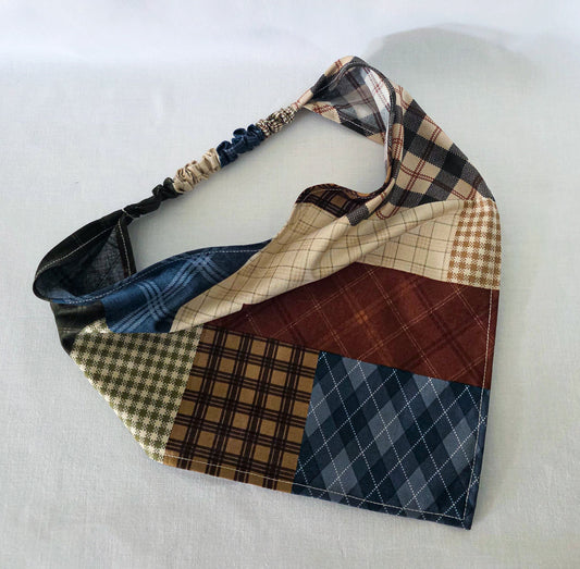 A handmade faux patchwork hair bandana with an elastic strap, featuring various plaid and checkered fabric panels in brown, beige, blue, and reddish brown, laid flat on a white fabric background.
