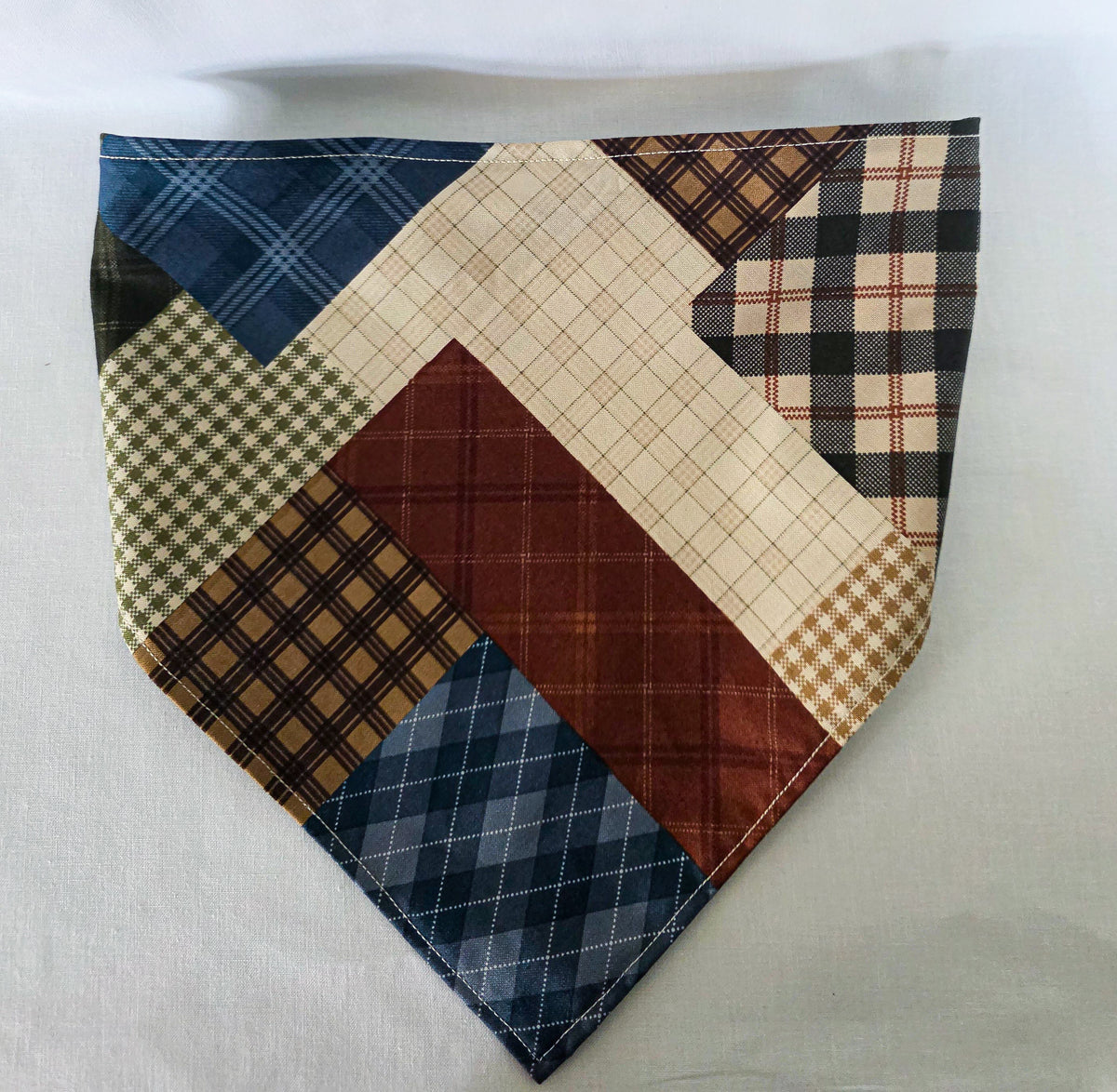 A handmade faux patchwork hair bandana with an elastic strap, featuring various plaid and checkered fabric panels in brown, beige, blue, and reddish brown, laid flat on a white fabric background.