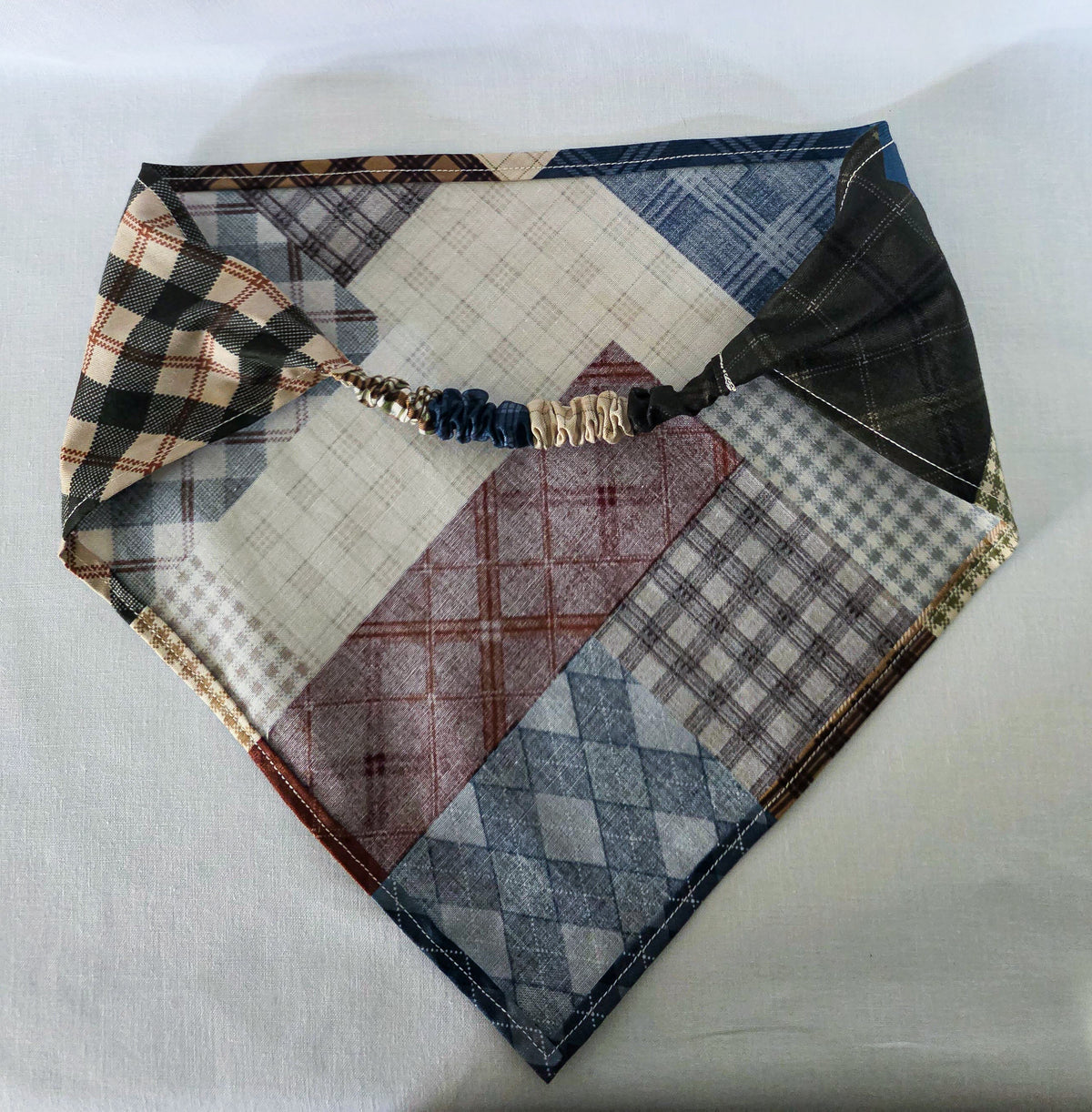 A handmade faux patchwork hair bandana with an elastic strap, featuring various plaid and checkered fabric panels in brown, beige, blue, and reddish brown, laid flat on a white fabric background. Its showing the backside of the bandana. 