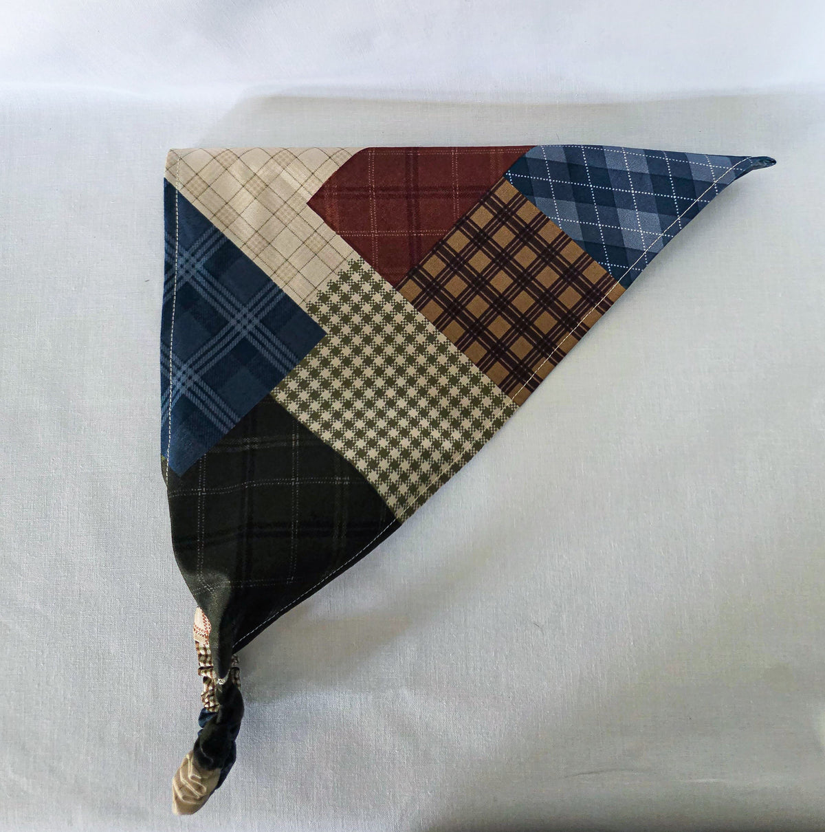 A handmade faux patchwork hair bandana with an elastic strap, featuring various plaid and checkered fabric panels in brown, beige, blue, and reddish brown, laid flat on its side on a white fabric background.