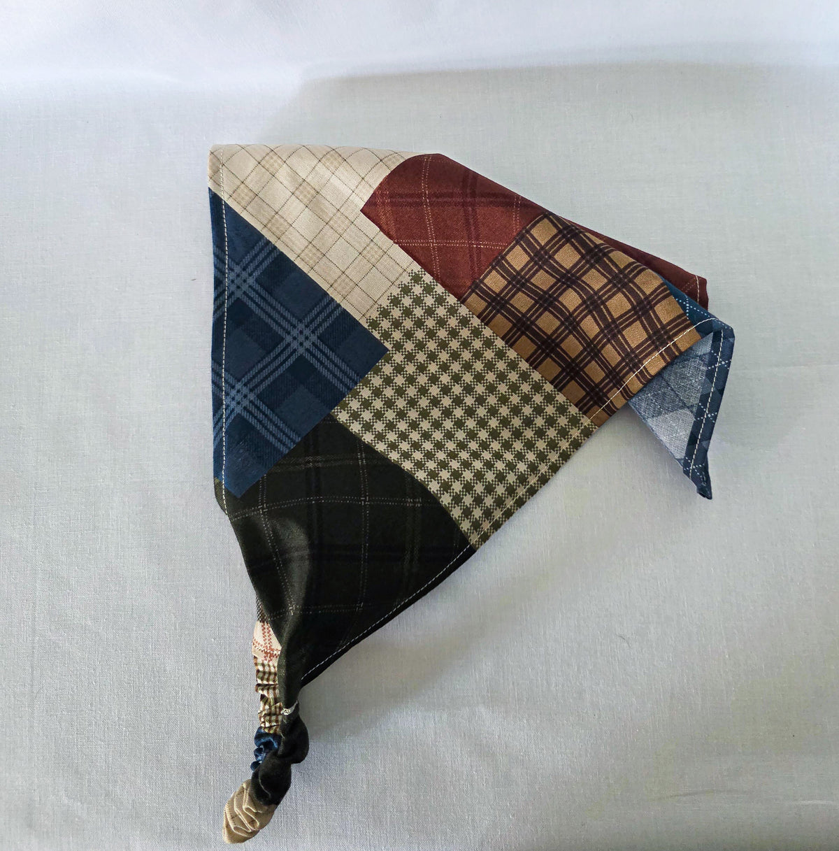 A handmade faux patchwork hair bandana with an elastic strap, featuring various plaid and checkered fabric panels in brown, beige, blue, and reddish brown, laid flat on its side on a white fabric background.
