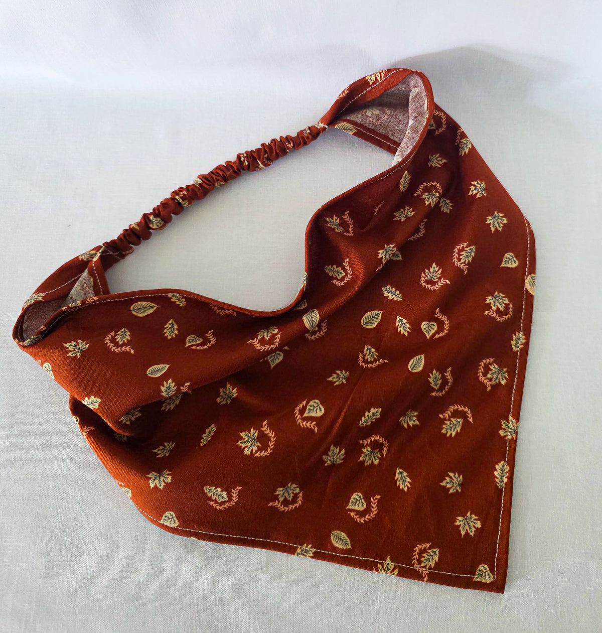 A burnt orange fabric bandana  with a small beige leaf pattern, featuring an elasticized strap, laid flat on a white background.