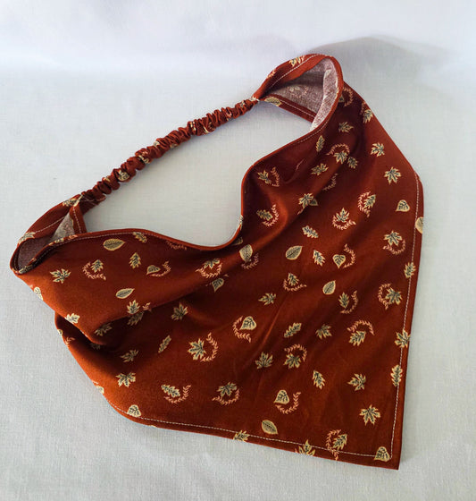 A burnt orange fabric bandana  with a small beige leaf pattern, featuring an elasticized strap, laid flat on a white background.