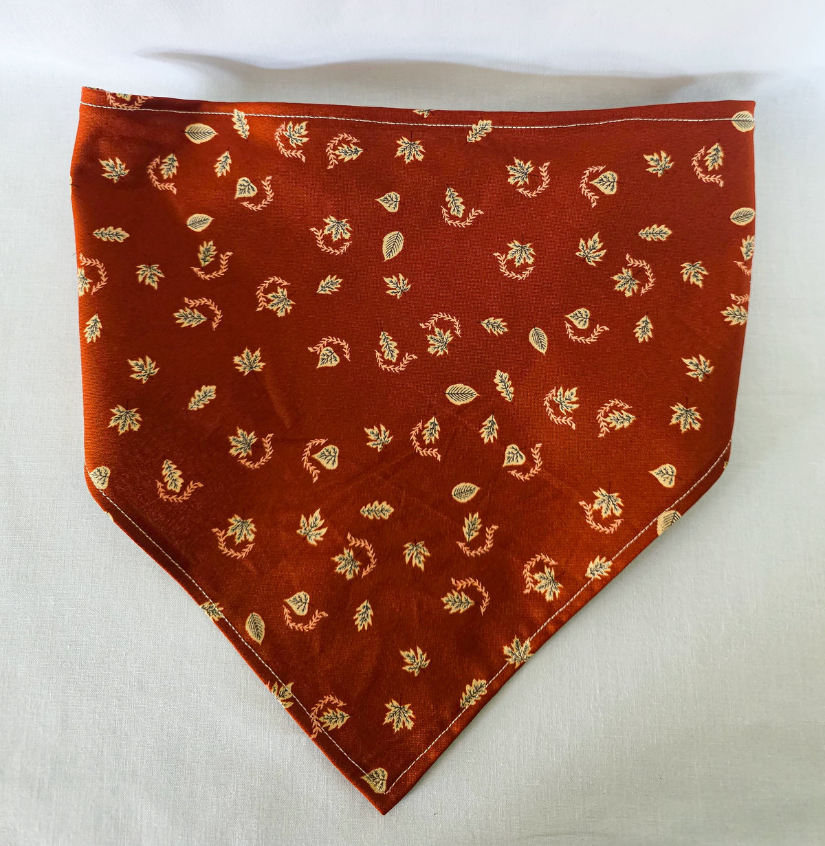 A burnt orange fabric bandana  with a small beige leaf pattern, featuring an elasticized strap, laid flat on a white background.
