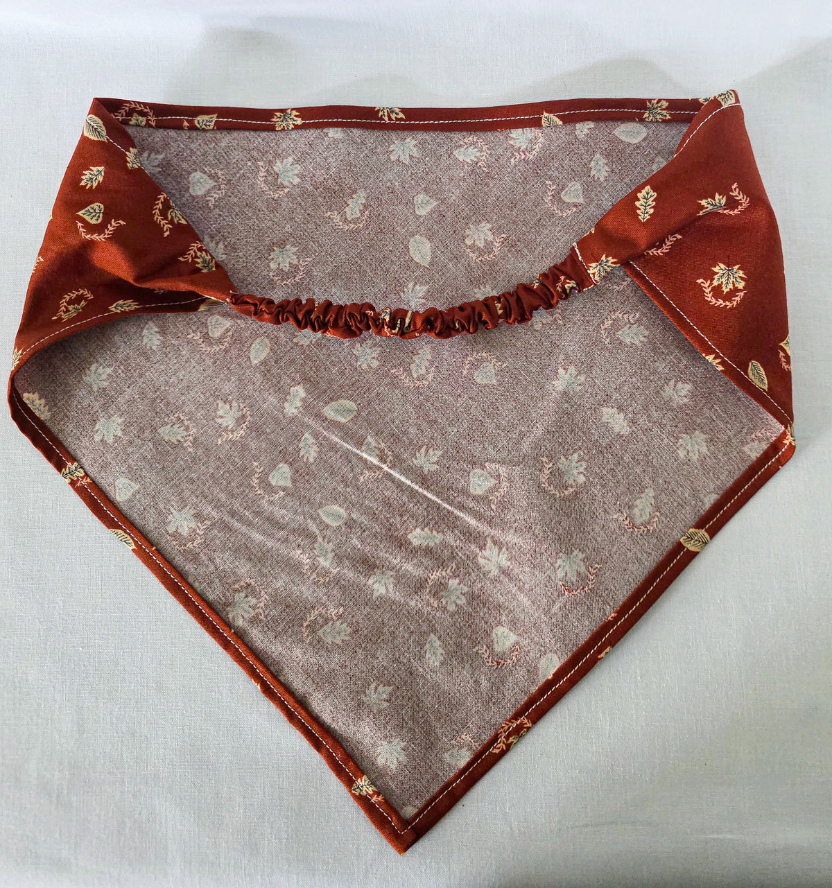 A burnt orange fabric bandana  with a small beige leaf pattern, featuring an elasticized strap, laid flat on a white background.