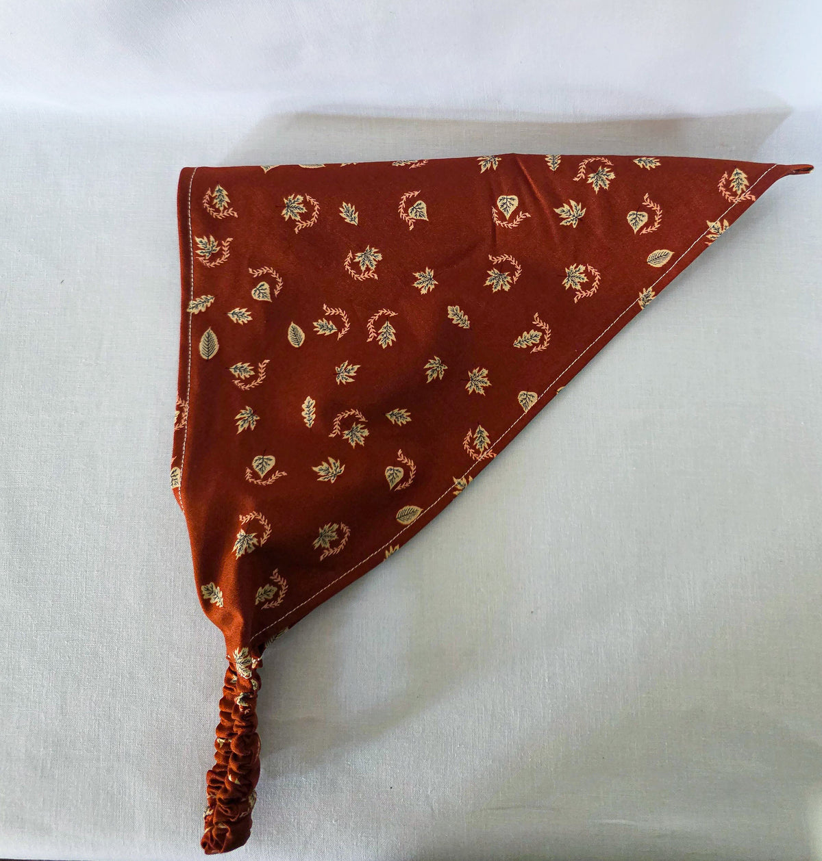 A burnt orange fabric bandana  with a small beige leaf pattern, featuring an elasticized strap, laid flat on its side on a white background.