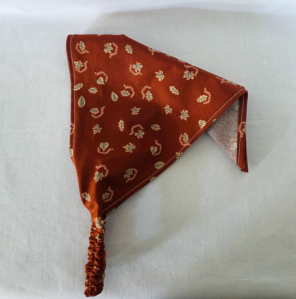 A burnt orange fabric bandana  with a small beige leaf pattern, featuring an elasticized strap, laid flat on its side on a white background.