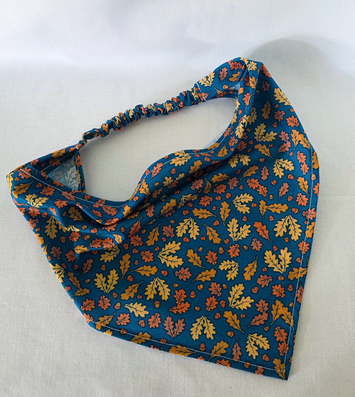 A triangular hair bandana with a gathered elastic strap, featuring an  autumn leaf pattern in yellow, orange, and brown with acorns on a dark teal blue background, laid flat on a white fabric surface.