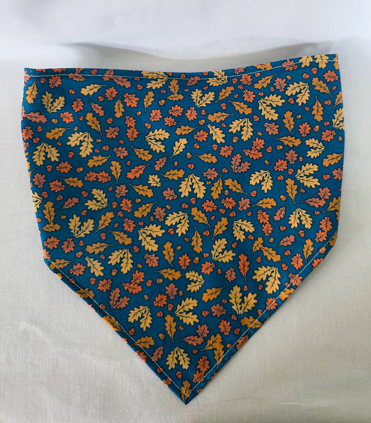 A triangular hair bandana with a gathered elastic strap, featuring an  autumn leaf pattern in yellow, orange, and brown with acorns on a dark teal blue background, laid flat on a white fabric surface.