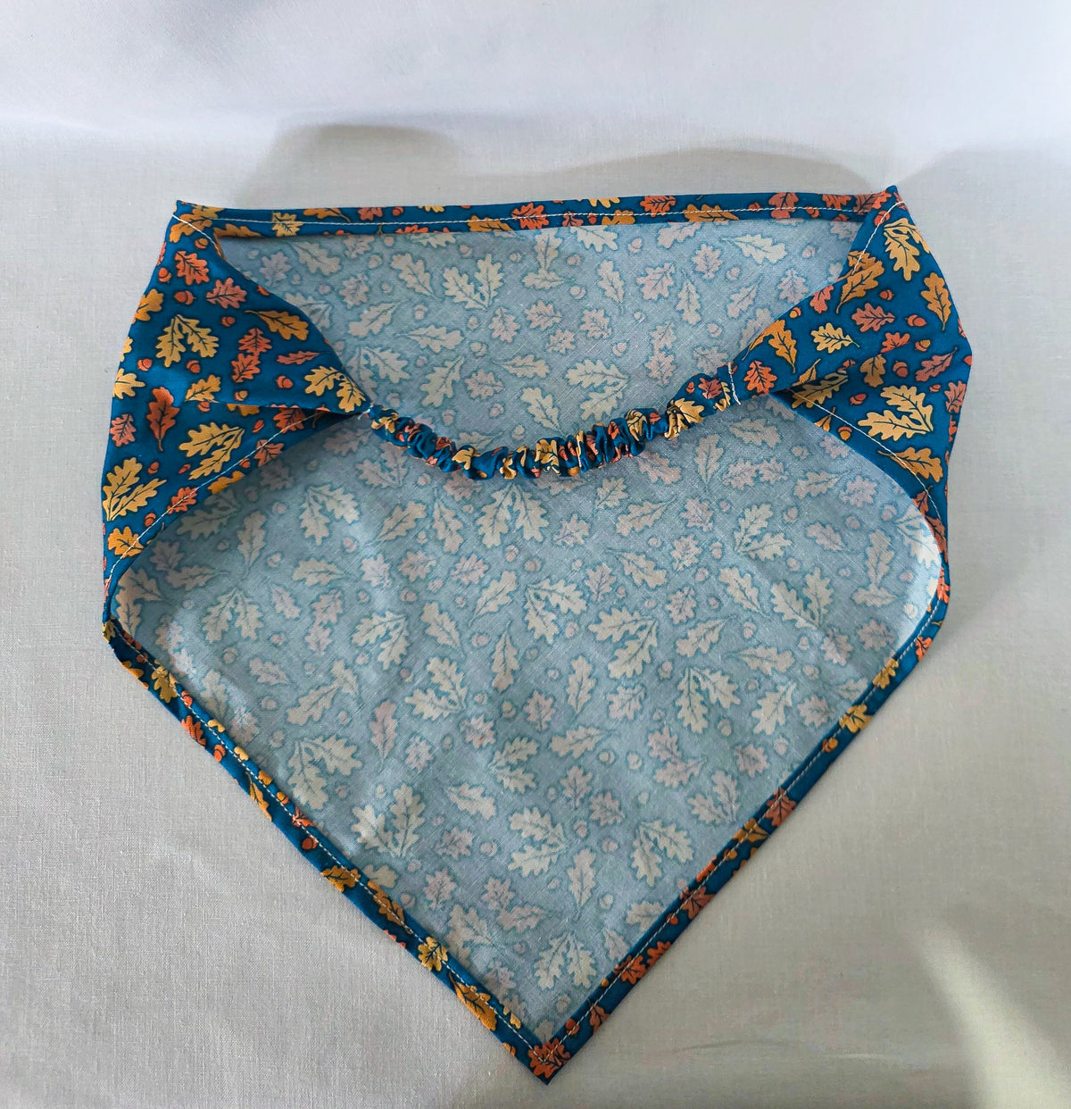 A triangular hair bandana with a gathered elastic strap, featuring an  autumn leaf pattern in yellow, orange, and brown with acorns on a dark teal blue background, laid flat on a white fabric surface.