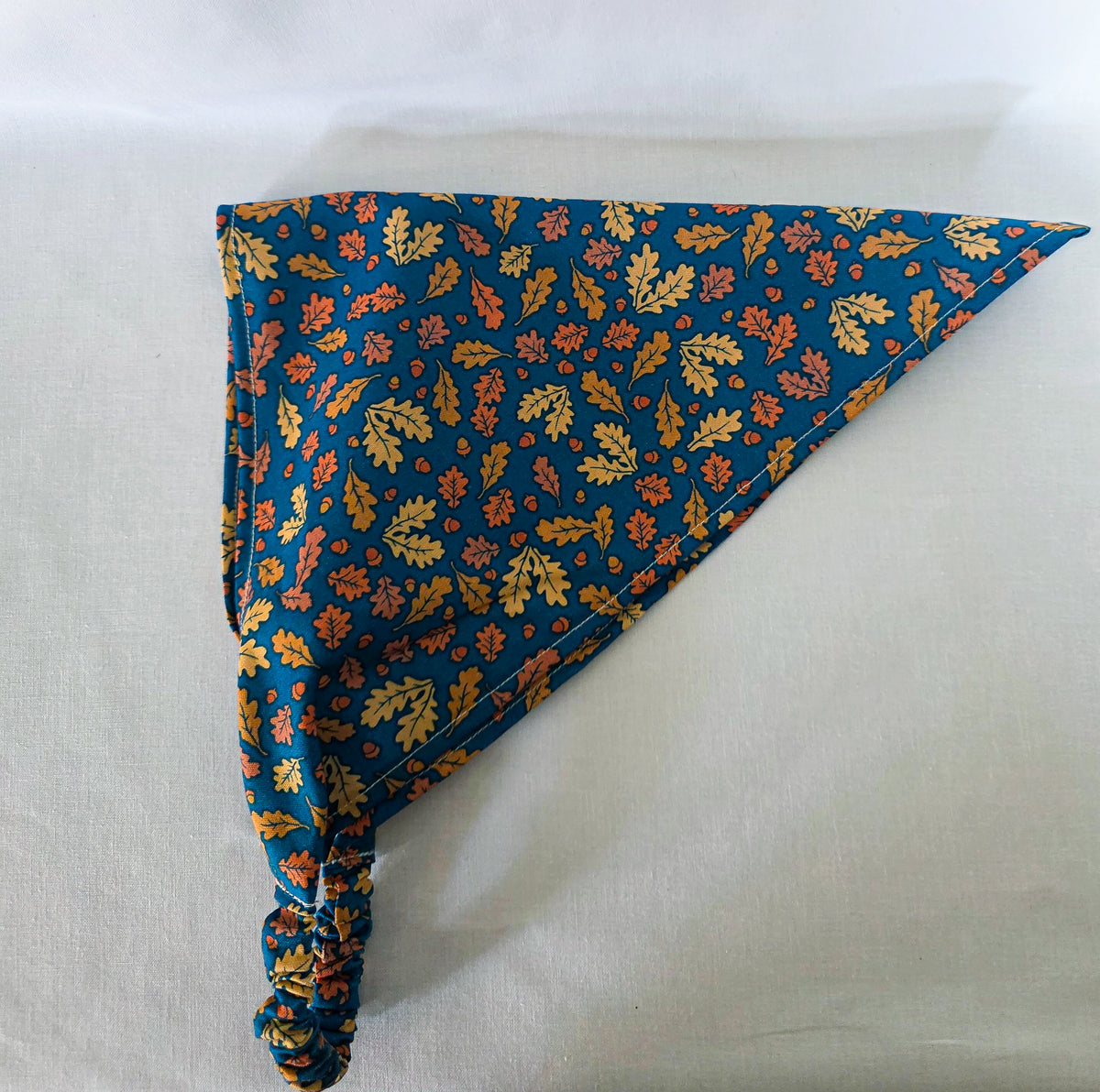 A triangular hair bandana with a gathered elastic strap, featuring an  autumn leaf pattern in yellow, orange, and brown with acorns on a dark teal blue background, laid flat on its side on a white fabric surface.
