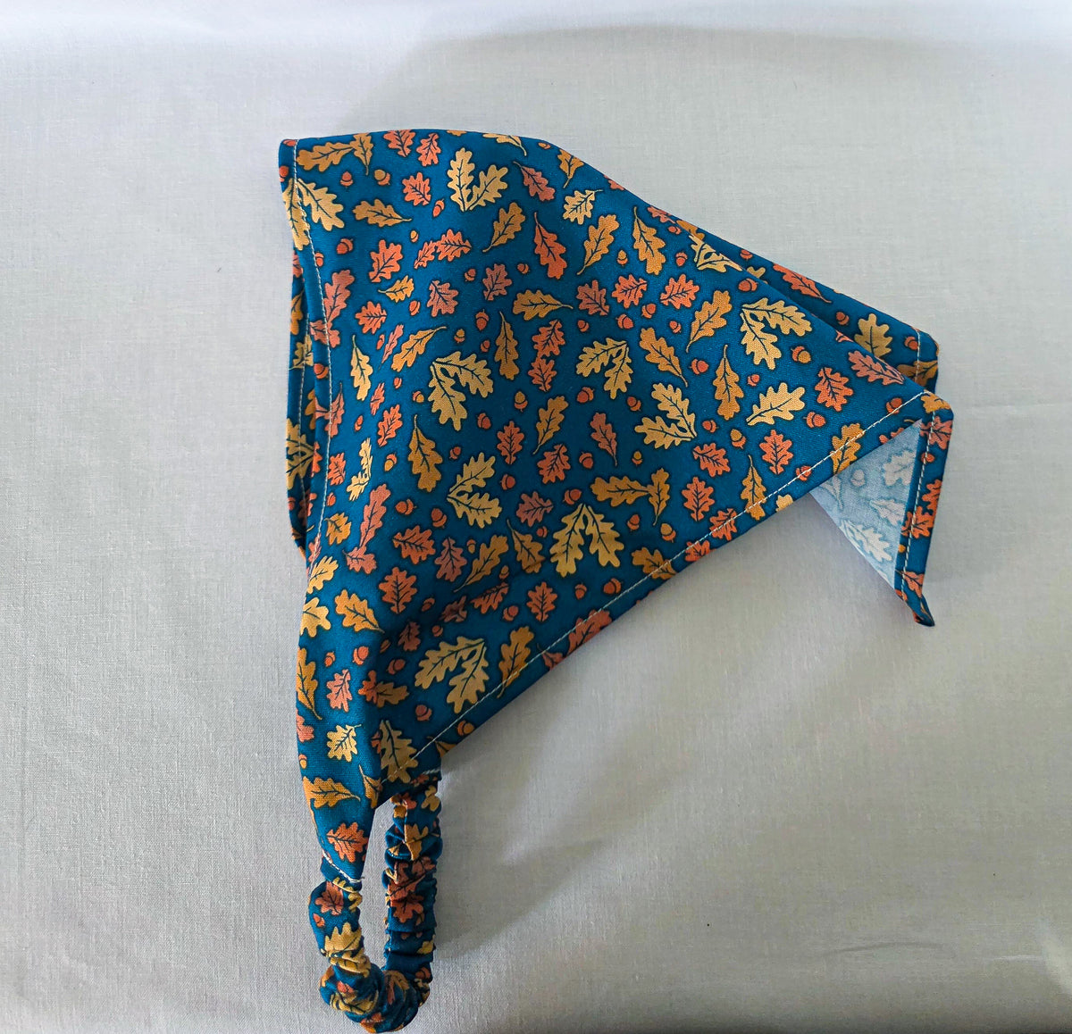 A triangular hair bandana with a gathered elastic strap, featuring an  autumn leaf pattern in yellow, orange, and brown with acorns on a dark teal blue background, laid flat on its side showing its drape on a white fabric surface.