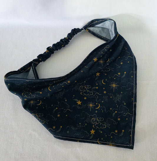 A dark navy blue hair bandana featuring a celestial print of gold stars, crescent moons, and clouds, with a matching elasticated strap. Laid on a white background. 