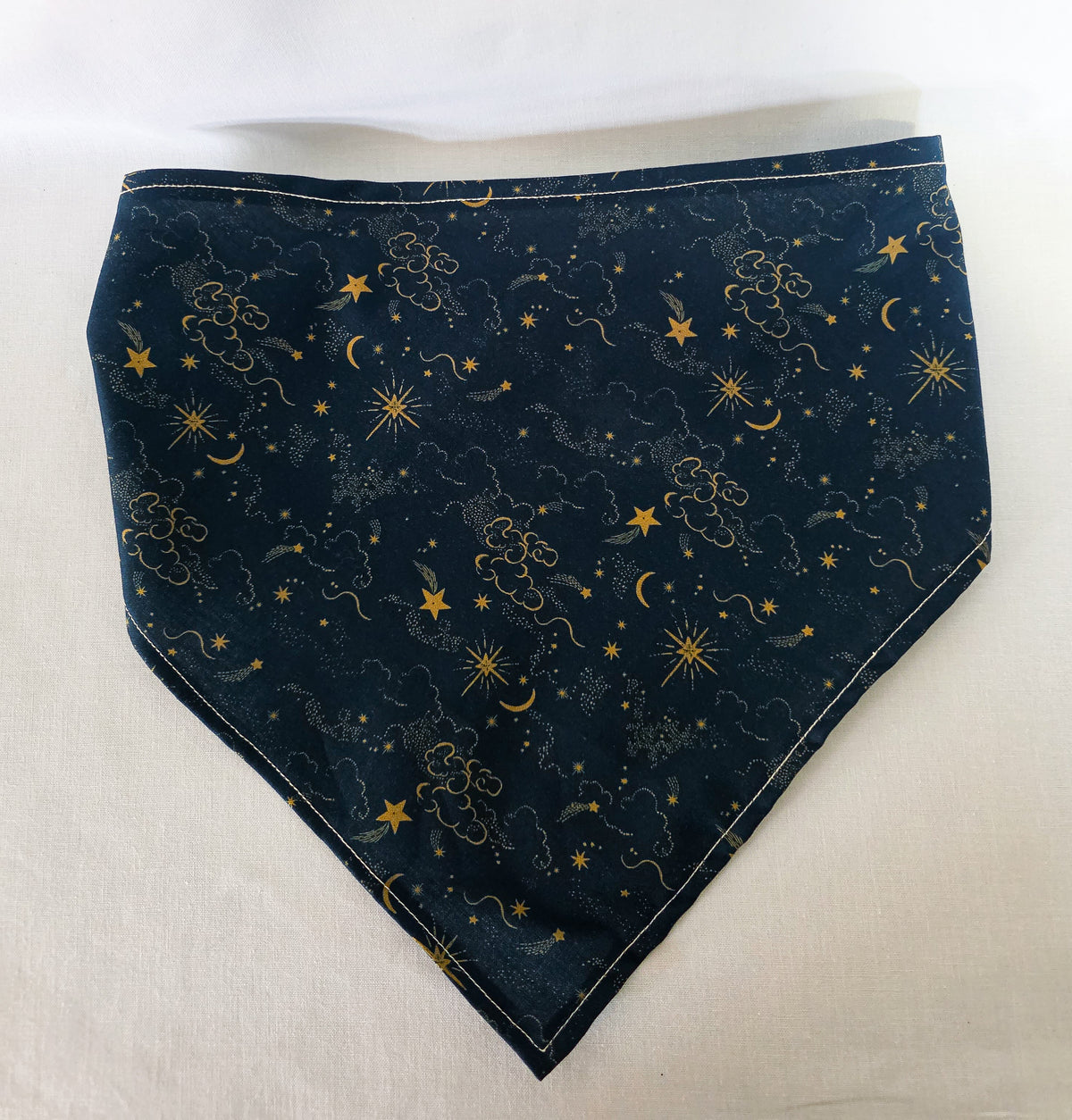 A dark navy blue hair bandana featuring a celestial print of gold stars, crescent moons, and clouds, with a matching elasticated strap. Laid flat on a white background. 