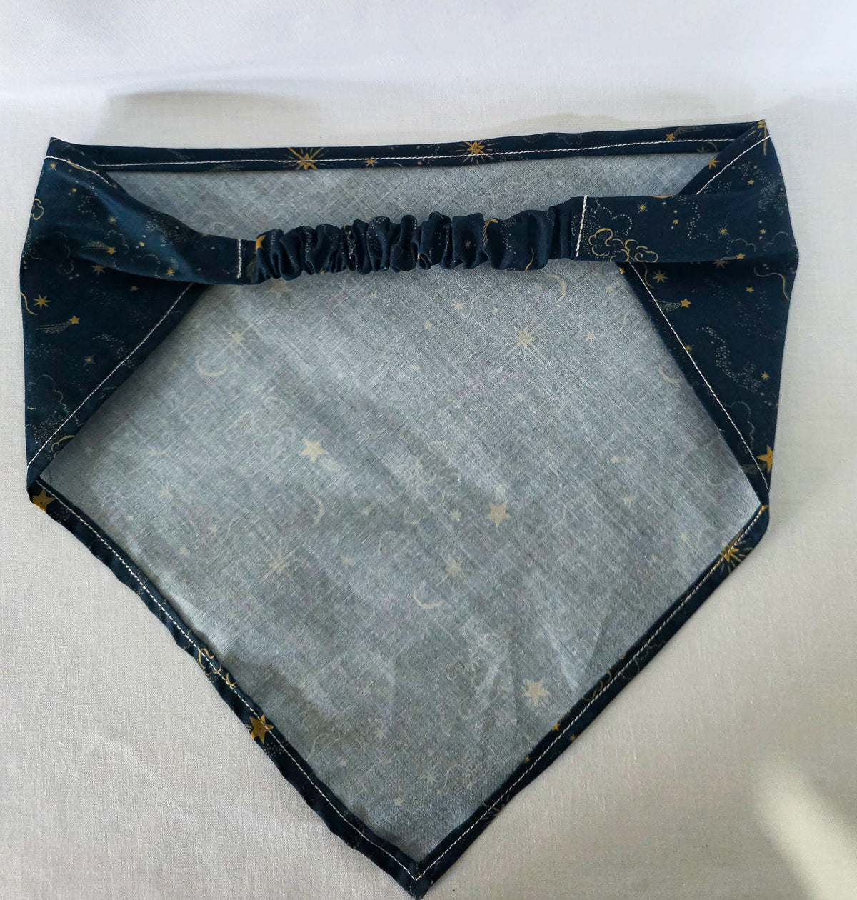A dark navy blue hair bandana featuring a celestial print of gold stars, crescent moons, and clouds, with a matching elasticated strap. Laid right side down, showing the backside, on a white background. 