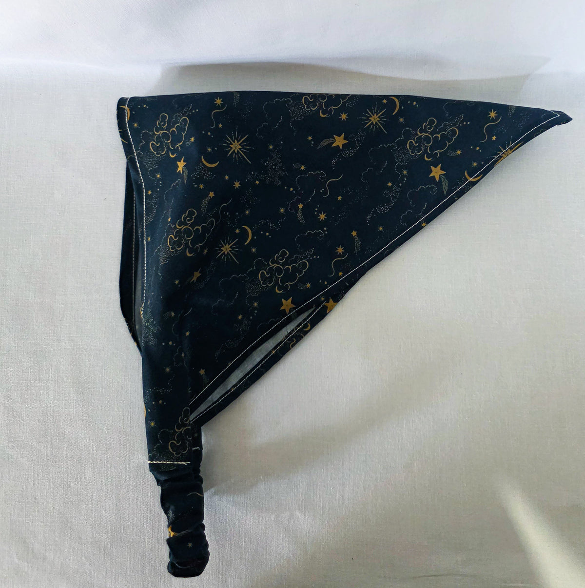 A dark navy blue hair bandana featuring a celestial print of gold stars, crescent moons, and clouds, with a matching elasticated strap. Laid on its side on a white background. 