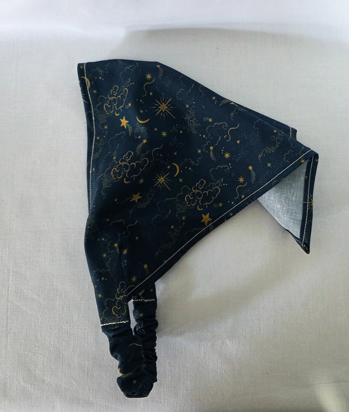A dark navy blue hair bandana featuring a celestial print of gold stars, crescent moons, and clouds, with a matching elasticated strap. Laid on its side a white background showing its drape. 