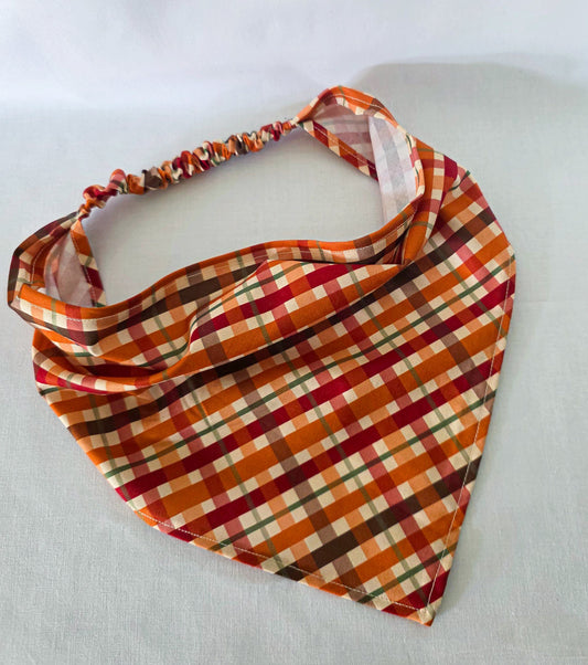 An elastic plaid hair bandana in orange, red, brown, and cream, laid on a white fabric background.