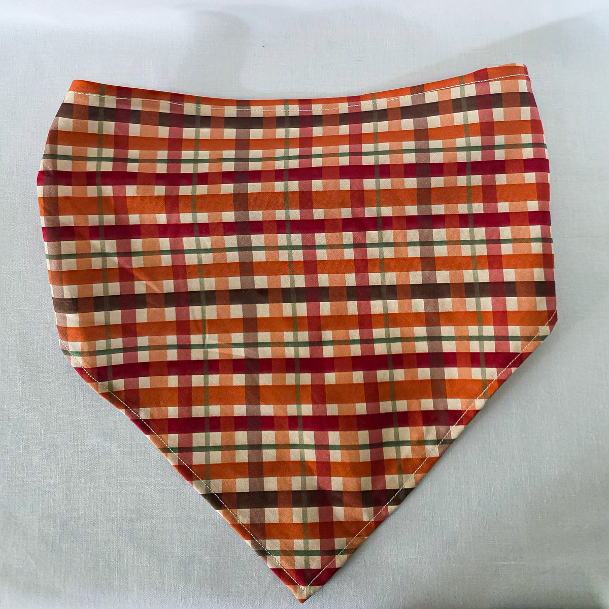 An elastic plaid hair bandana in orange, red, brown, and cream, laid flat on a white fabric background.