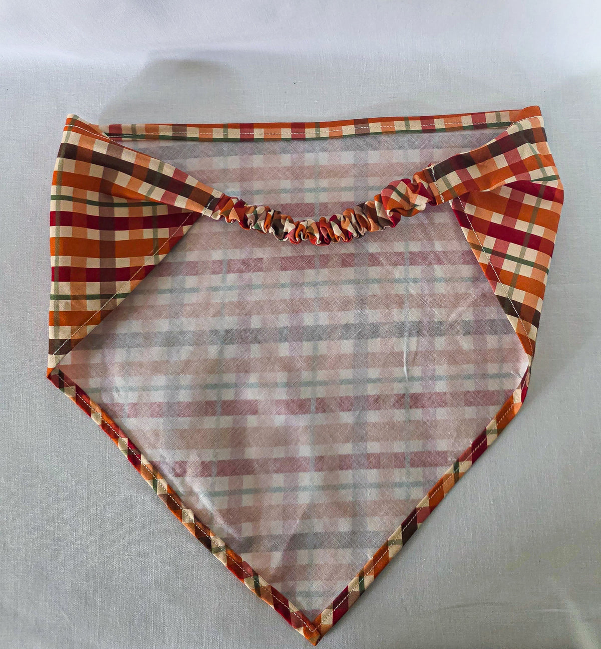 An elastic plaid hair bandana in orange, red, brown, and cream, laid right side down on a white fabric background, showing the elastic band.