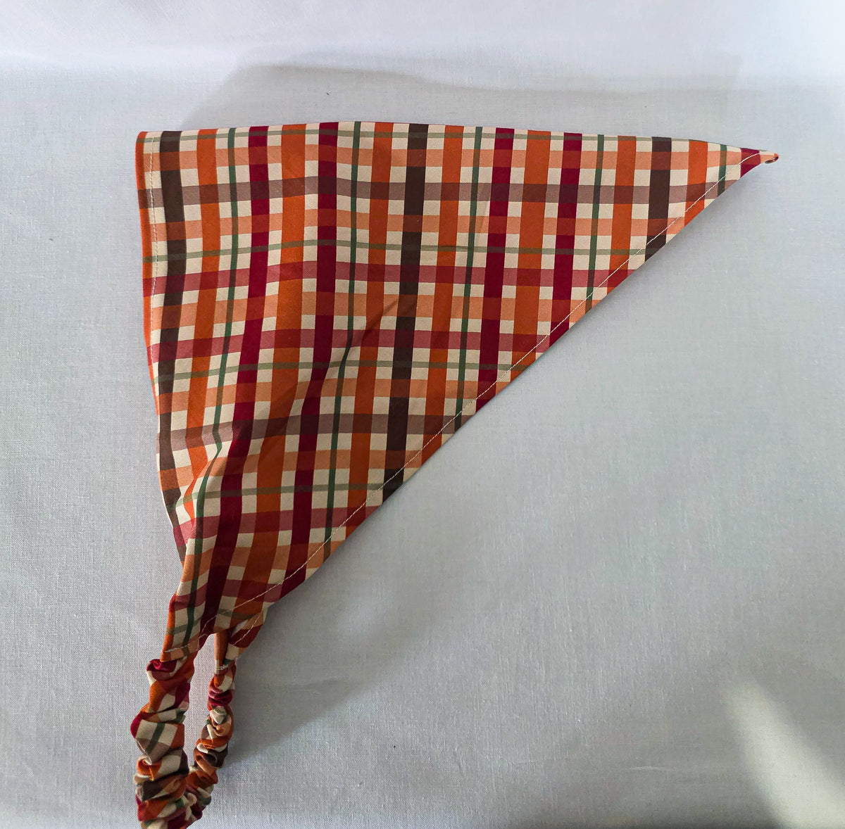 An elastic plaid hair bandana in orange, red, brown, and cream, laid on its side on a white fabric background.