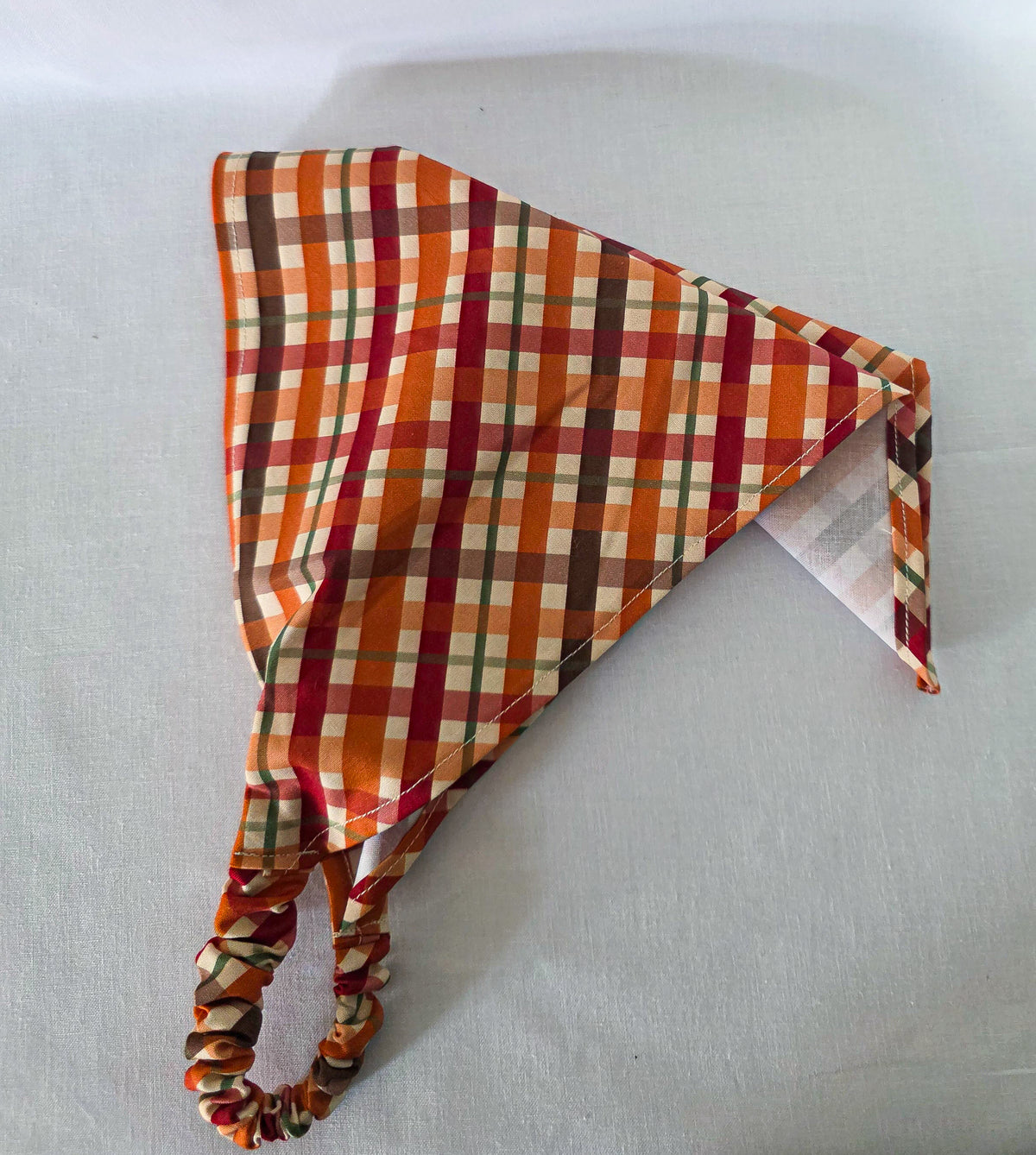 An elastic plaid hair bandana in orange, red, brown, and cream, laid on its side showing its drape on a white fabric background.
