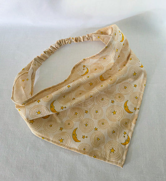 An elastic hair bandana with a yellow/gold celestial moon and star print throughout. Laid on a white fabric background.  