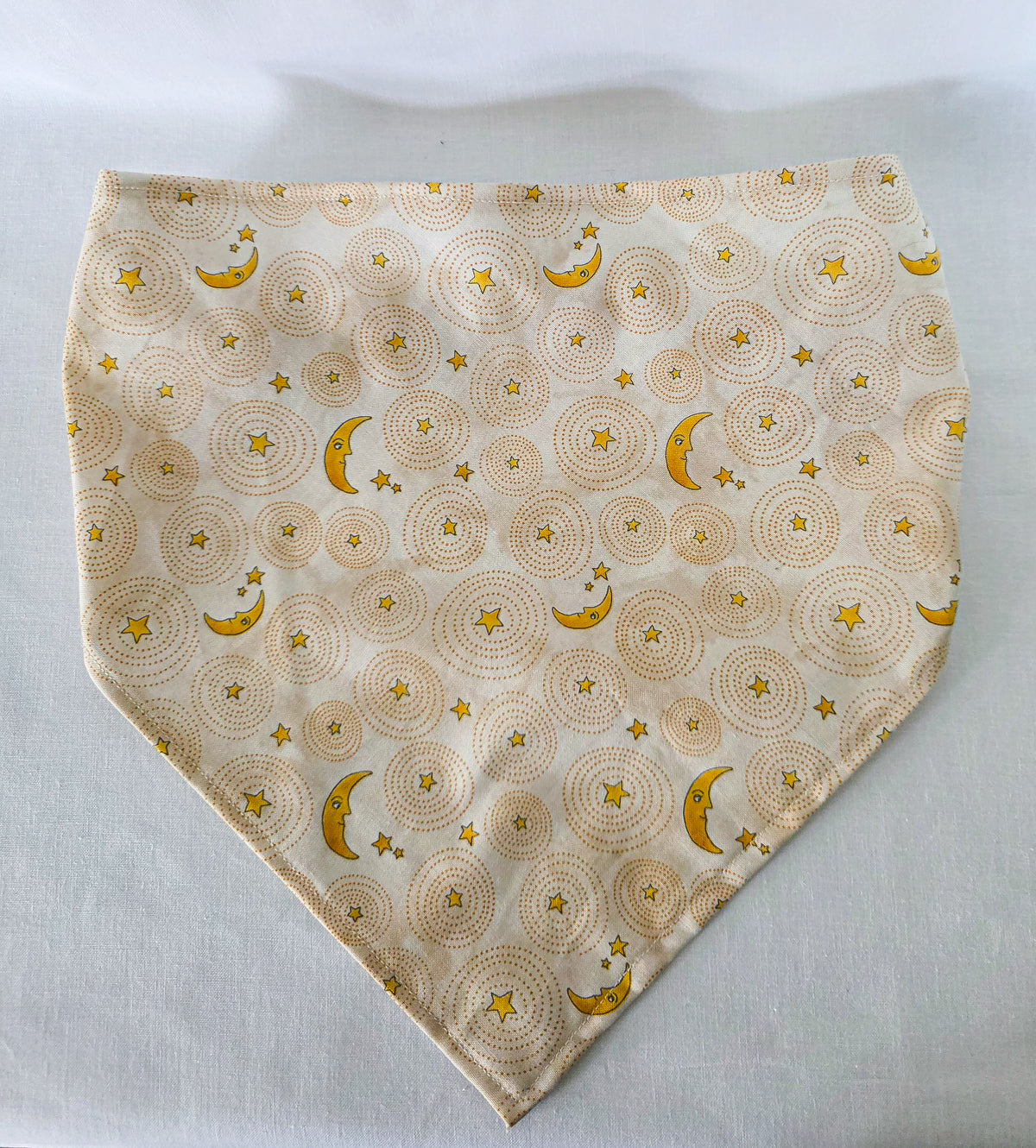 A triangular hair bandana made of cream-colored fabric with a repeating pattern of yellow crescent moons and stars over circular dotted sunburst designs, laid flat on a white background.