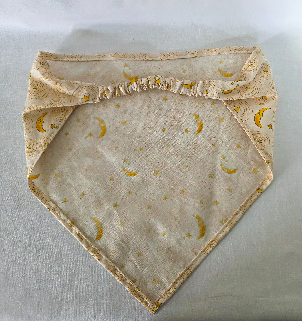 A triangular hair bandana made of cream-colored fabric with a repeating pattern of yellow crescent moons and stars over circular dotted sunburst designs, laid flat on a white background.