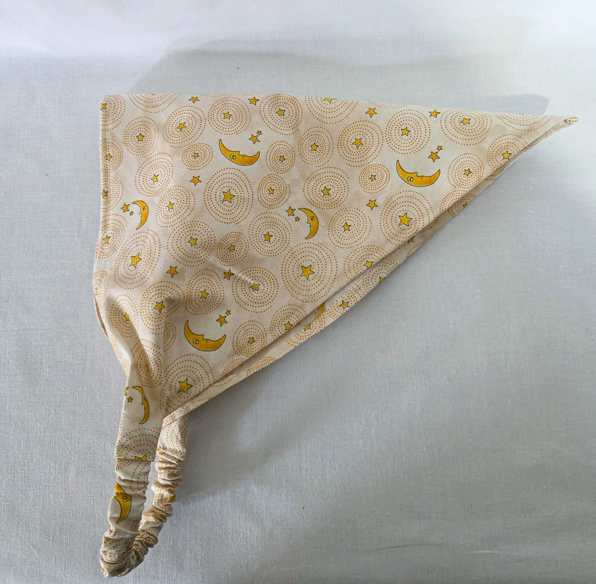 A triangular hair bandana made of cream-colored fabric with a repeating pattern of yellow crescent moons and stars over circular dotted sunburst designs, laid flat on its side on a white background.