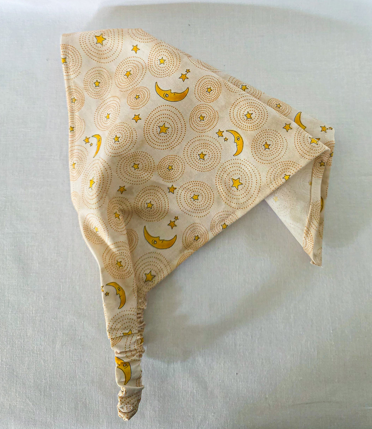 A triangular hair bandana made of cream-colored fabric with a repeating pattern of yellow crescent moons and stars over circular dotted sunburst designs, laid flat on its side showing the drape on a white background.