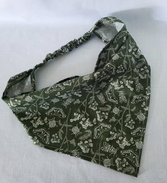 A green elastic hair bandana with a white floral and bug print on it. Laid on a white fabric background. 